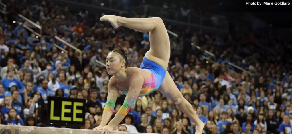 Emily Lee holds a pose on balance beam. She leans over her hands clutching the beam, with her right foot down on the beam and her left leg bent over her head and her toe pointed.
